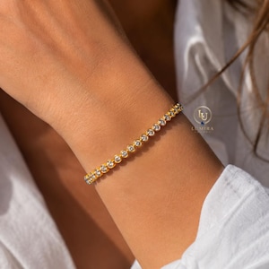 May include: A gold bracelet featuring a line of clear, round gemstones. The bracelet is worn on a wrist, against a white shirt. This elegant piece of jewellery is a delicate accessory.