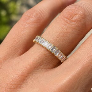 Peut inclure: Une bague en or avec une rangée continue de pierres transparentes rectangulaires taille baguette, fixées par des griffes partagées. Les pierres sont disposées horizontalement sur toute la circonférence de l'anneau en or jaune poli.