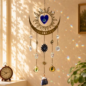 May include: A hanging suncatcher featuring a gold-toned metal crescent moon and sun frame. It includes a blue heart-shaped evil eye charm, a dark blue flower-shaped evil eye, dangling chains with clear crystals, small eye charms, and a raw blue stone.