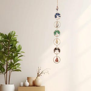 May include: A vertical hanging ornament featuring six circular gold-toned metal frames connected by small rings. The frames contain wire-wrapped tree sculptures with blue, green, and brown gemstone chips, alternating with raw crystal pendants.