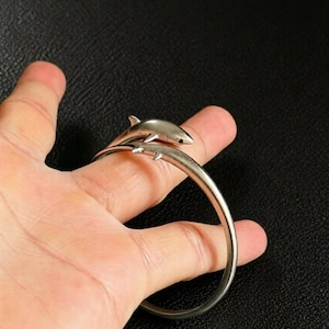 Puede incluir: Una pulsera de brazalete de metal en tono plateado con un diseño de tiburón esculpido. El tiburón está colocado a lo largo de la parte superior de la banda, con su cuerpo curvándose para seguir la forma de la pulsera. El metal tiene un acabado liso.