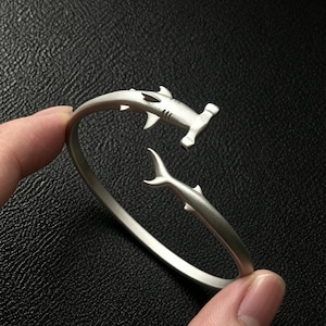 Puede incluir: Un brazalete abierto de tono plateado con un diseño de tiburón martillo. La pulsera tiene un acabado mate, con la cabeza del tiburón formando un extremo y la cola el otro, creando una apariencia envolvente y continua.