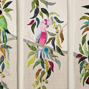 May include: A textile wall hanging featuring a central panel with a pink and grey cockatoo perched on a branch surrounded by multicolored fabric leaves in shades of green, teal, and magenta against a textured cream-colored quilted background.
