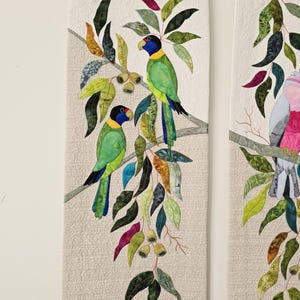Op de afbeelding: Een verticale stoffen wandhanger met een gewatteerde crèmekleurige achtergrond en een applicatieontwerp van twee groene papegaaien met zwarte koppen en gele kragen, zittend op een tak omringd door veelkleurige eucalyptusbladeren en zaaddozen.