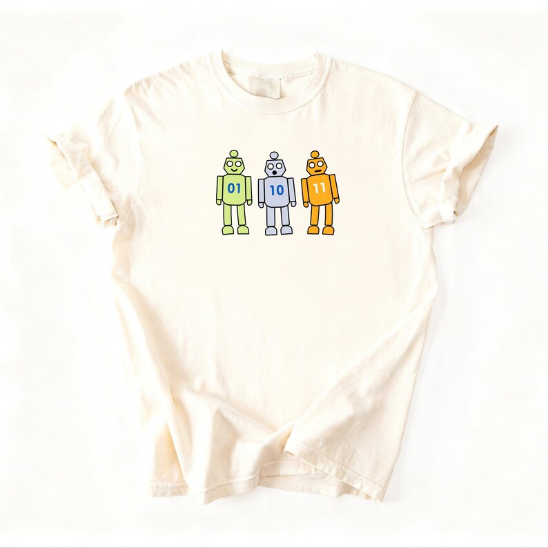 May include: A cream-colored short-sleeve t-shirt featuring a graphic of three cartoon robots in a row. The left robot is light green with the number 01, the middle robot is light blue with 10, and the right robot is orange with the number 11.