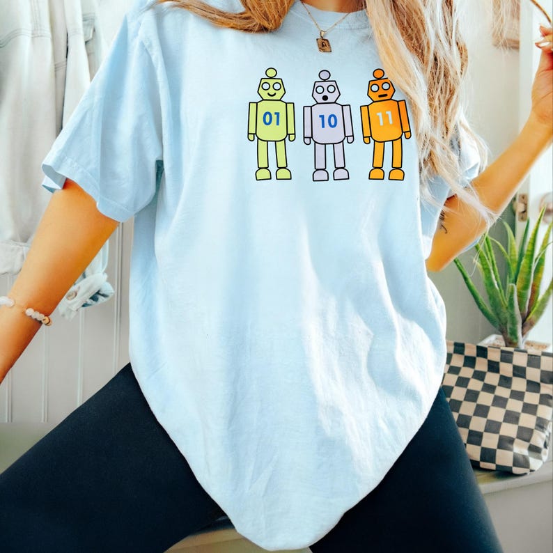 May include: A light blue t-shirt featuring a graphic of three cartoon robots in lime green, light purple, and orange. Each robot has a binary number printed on its chest: 01 on the green robot, 10 on the purple robot, and 11 on the orange robot.