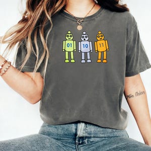 May include: A charcoal gray short-sleeve t-shirt featuring a graphic of three cartoon robots in light green, light blue, and orange. Each robot displays a binary number on its chest: 01, 10, and 11 respectively. The shirt has a crew neckline.