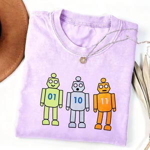 May include: A light purple crew neck t-shirt featuring a graphic of three cartoon robots in lime green, silver, and orange. The robots have blue binary numbers 01, 10, and 11 printed on their chests. The shirt has a ribbed collar and short sleeves.