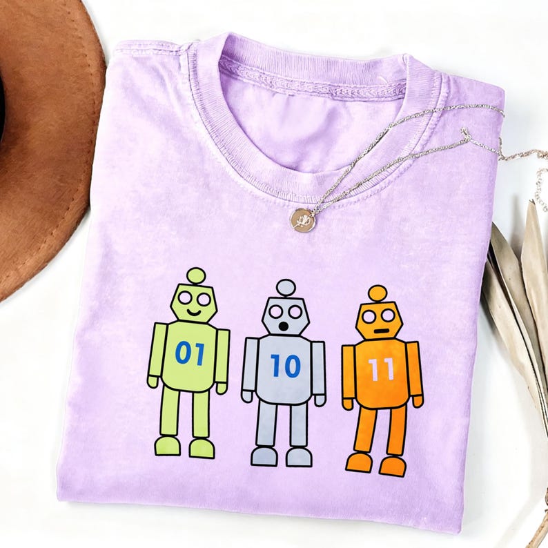 May include: A light purple crew neck t-shirt featuring a graphic of three cartoon robots in lime green, silver, and orange. The robots have blue binary numbers 01, 10, and 11 printed on their chests. The shirt has a ribbed collar and short sleeves.