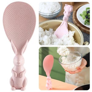 Puede incluir: Una paleta de arroz rosa con un mango en forma de conejito. La paleta tiene una superficie texturizada y se muestra sacando arroz de una olla y un cuenco. El mango de conejito añade un toque caprichoso al utensilio de cocina.