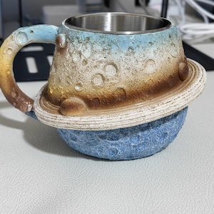 Peut inclure: Une tasse avec un intérieur métallique et un extérieur texturé en forme de planète avec un anneau. La partie supérieure présente une surface cratérisée dans des tons de bleu, blanc et marron, tandis que la partie inférieure est une sphère bleue.