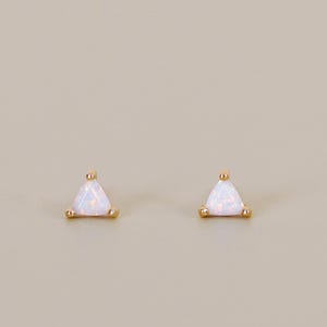 May include: A pair of gold-colored stud earrings, each featuring a triangular, iridescent opal stone. The earrings have a simple, minimalist design, with the opal stones set in a gold-tone metal. The studs are small and delicate, suitable for everyday wear.