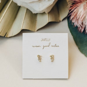 May include: A pair of gold earrings with small, clear baguette-shaped stones. The earrings are displayed on a white card with the text "Jaxkelly wear good vibes".