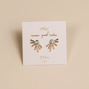 May include: A pair of gold-colored stud earrings with an opal center stone and a sunburst design. The earrings are displayed on a white card with the text "JAXKELLY wear good vibes OPAL joy".