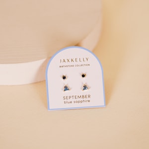 May include: A set of three gold earrings with blue sapphire stones. The earrings are on a white card with the text "JAXKELLY BIRTHSTONE COLLECTION SEPTEMBER blue sapphire".