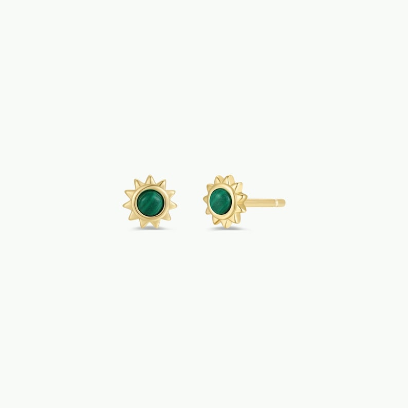May include: A pair of gold-coloured stud earrings. Each earring features a sunburst design with a central, round, green stone. The earrings are displayed on a white background.