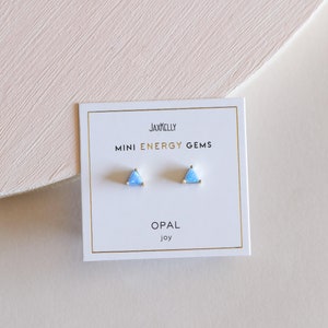 May include: A pair of small triangle-shaped earrings with blue opal stones. The earrings are on a white card with the text "JAXKELLY MINI ENERGY GEMS OPAL joy".