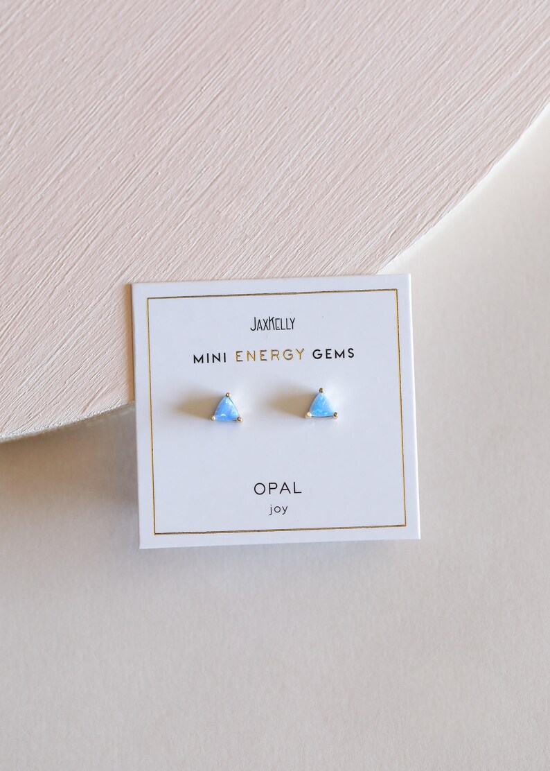 May include: A pair of small triangle-shaped earrings with blue opal stones. The earrings are on a white card with the text "JAXKELLY MINI ENERGY GEMS OPAL joy".