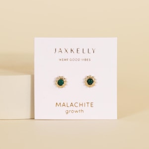 May include: Gold-coloured stud earrings featuring a malachite stone centre and a sunburst design. The earrings are presented on a white card with the text "JAXKELLY" and "WEAR GOOD VIBES". The card also reads "MALACHITE growth".