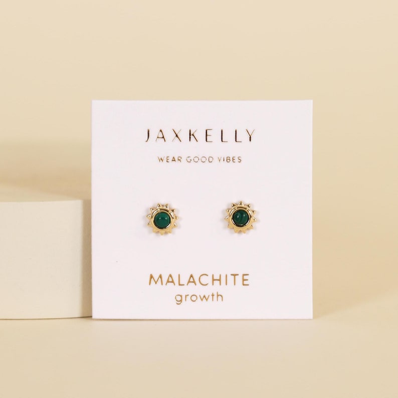 May include: Gold-coloured stud earrings featuring a malachite stone centre and a sunburst design. The earrings are presented on a white card with the text "JAXKELLY" and "WEAR GOOD VIBES". The card also reads "MALACHITE growth".