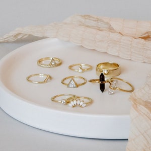 Puede incluir: Una colecci&oacute;n de anillos de oro con diferentes piedras preciosas y dise&ntilde;os, incluyendo un anillo con una piedra en forma de tri&aacute;ngulo, un anillo con una fila de piedras peque&ntilde;as y un anillo con un grupo de piedras blancas. Los anillos est&aacute;n dispuestos en un plato blanco.