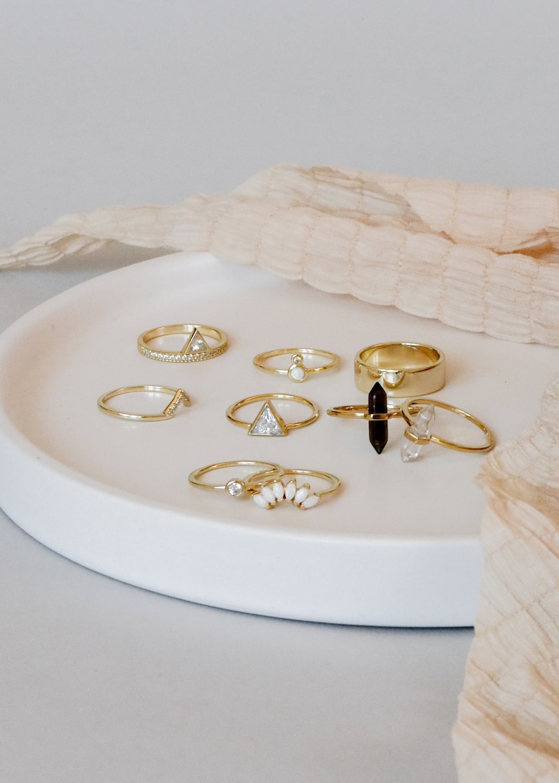 Puede incluir: Una colecci&oacute;n de anillos de oro con diferentes piedras preciosas y dise&ntilde;os, incluyendo un anillo con una piedra en forma de tri&aacute;ngulo, un anillo con una fila de piedras peque&ntilde;as y un anillo con un grupo de piedras blancas. Los anillos est&aacute;n dispuestos en un plato blanco.