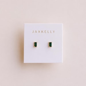 May include: A pair of gold stud earrings featuring rectangular emerald green stones, mounted on a white display card with the text JAXKELLY printed in gold lettering at the top center. The earrings are positioned side by side on the card.