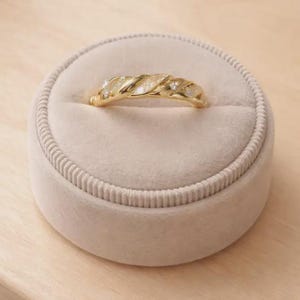 May include: A gold-colored ring with a twisted rope design, set with small, sparkling stones. The ring is displayed inside a round, beige velvet ring box, set on a light wood surface. The ring's design is a continuous, textured pattern.