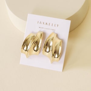 May include: A pair of gold-colored earrings with a wavy design, displayed on a white card. The card features the text "JAXKELLY" above "WEAR GOOD VIBES" in a simple font. The earrings have a polished, reflective surface.