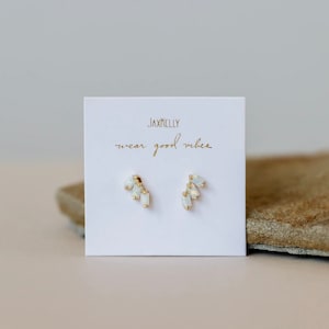 May include: A pair of gold earrings with white opal stones. The earrings are displayed on a white card with the text "Jaxkelly wear good vibes".