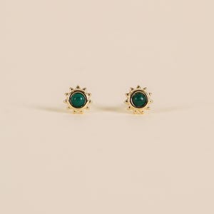 May include: A pair of gold-coloured stud earrings. Each earring has a dark green, circular stone at the centre, surrounded by a sunburst design. The earrings are set against a neutral background.