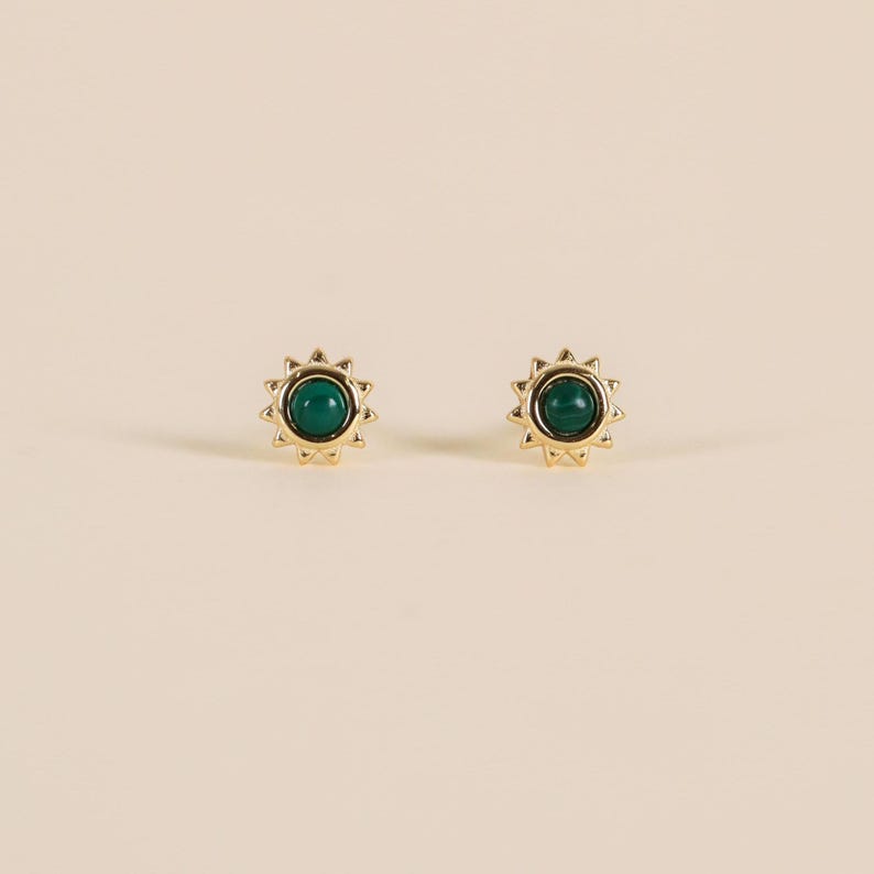 May include: A pair of gold-coloured stud earrings. Each earring has a dark green, circular stone at the centre, surrounded by a sunburst design. The earrings are set against a neutral background.