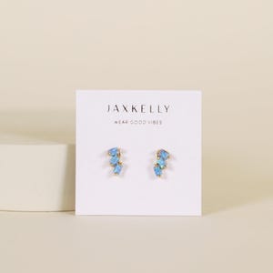 May include: A pair of gold-toned stud earrings with light blue, irregular-shaped stones. The earrings are displayed on a white card with the text "JAXKELLY" and "WEAR GOOD VIBES". The background is a soft, neutral color.