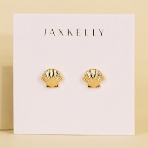 May include: A pair of gold-toned stud earrings shaped like small seashells, mounted on a white rectangular display card. The text JAXKELLY is printed in black, centered at the top of the card above the two identical shell-shaped earrings.