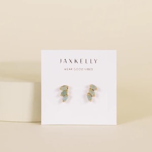 May include: A pair of gold-toned stud earrings with three light green, irregular-shaped stones. The earrings are displayed on a white card with the text "JAXKELLY" and "WEAR GOOD VIBES".