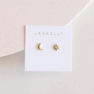 May include: A pair of gold stud earrings on a white display card with the text JAXKELLY. One earring is a crescent moon shape with a small embedded stone, and the other is a starburst shape with a central stone. Both earrings are metallic gold.