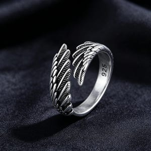 May include: A silver ring with an open design, featuring wing-like details. The ring has a textured surface with feather-like patterns. The number "925" is visible on the ring's band. The ring is set against a dark background.