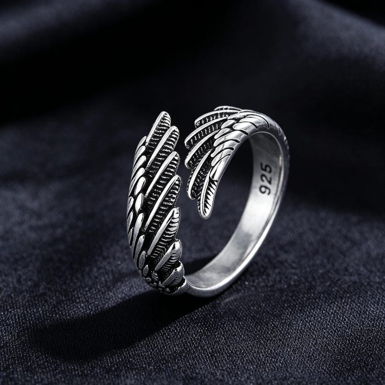 May include: A silver ring with an open design, featuring wing-like details. The ring has a textured surface with feather-like patterns. The number "925" is visible on the ring's band. The ring is set against a dark background.