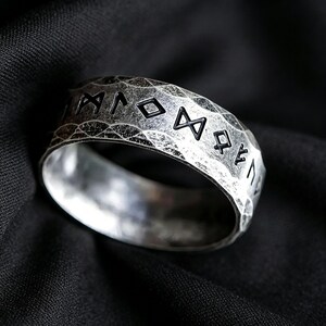 Peut inclure: Une bague argent&eacute;e avec une surface martel&eacute;e et textur&eacute;e pr&eacute;sentant des symboles runiques noirs grav&eacute;s. L'anneau a un profil large et plat avec une finition vieillie et us&eacute;e qui met en valeur les lettres sombres en creux sur l'ext&eacute;rieur.