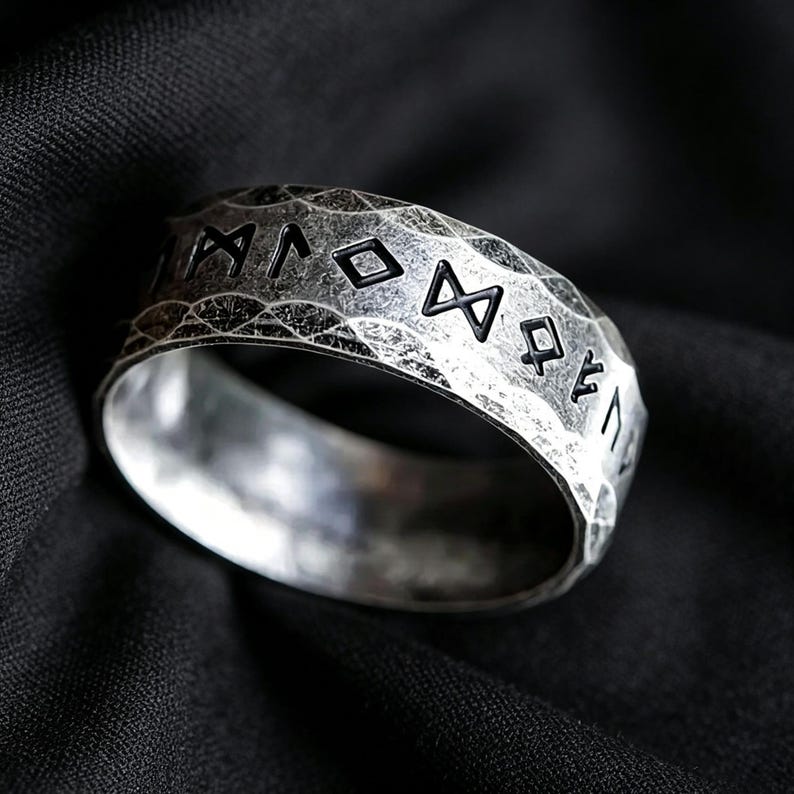 Peut inclure: Une bague argent&eacute;e avec une surface martel&eacute;e et textur&eacute;e pr&eacute;sentant des symboles runiques noirs grav&eacute;s. L'anneau a un profil large et plat avec une finition vieillie et us&eacute;e qui met en valeur les lettres sombres en creux sur l'ext&eacute;rieur.