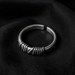 Peut inclure: Une bague ouverte de couleur argent&eacute;e pr&eacute;sentant un anneau textur&eacute; avec une s&eacute;rie de d&eacute;tails ressemblant &agrave; du fil enroul&eacute; sur le devant. L'int&eacute;rieur de l'anneau est clairement marqu&eacute; du texte S925, indiquant la composition de ce bijou r&eacute;glable.