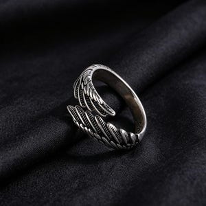 May include: A silver ring with a detailed feather design. The ring wraps around the finger with an open design, featuring intricate feather detailing. The ring is set against a dark background.