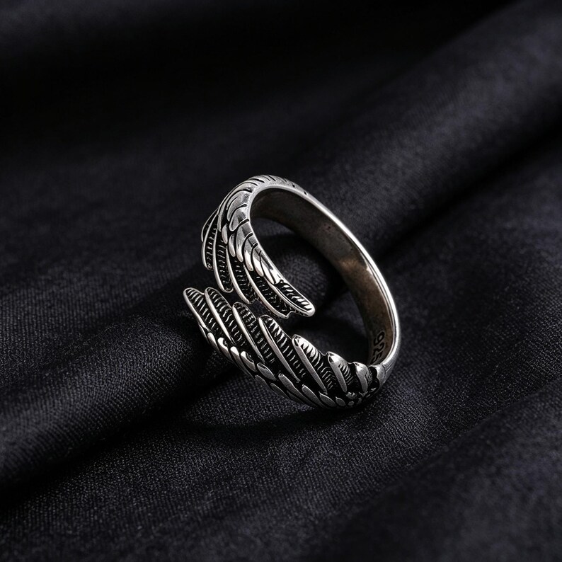 May include: A silver ring with a detailed feather design. The ring wraps around the finger with an open design, featuring intricate feather detailing. The ring is set against a dark background.