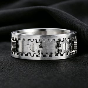 May include: A silver-toned metal ring featuring a wide band with a brushed finish. The exterior surface is decorated with a series of small, raised gear-shaped elements that are evenly spaced around the circumference of the band.