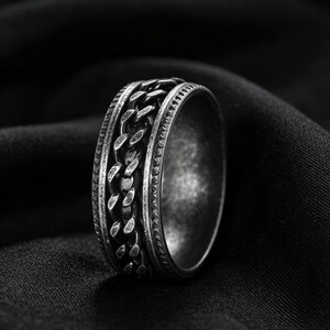 Peut inclure: Une bague en m&eacute;tal argent&eacute; dot&eacute;e d'une bande centrale rotative &agrave; maillons de cha&icirc;ne, encadr&eacute;e par deux bords textur&eacute;s avec une finition sombre et oxyd&eacute;e. L'anneau pr&eacute;sente un profil large et robuste &agrave; l'aspect vieilli et d&eacute;taill&eacute;.