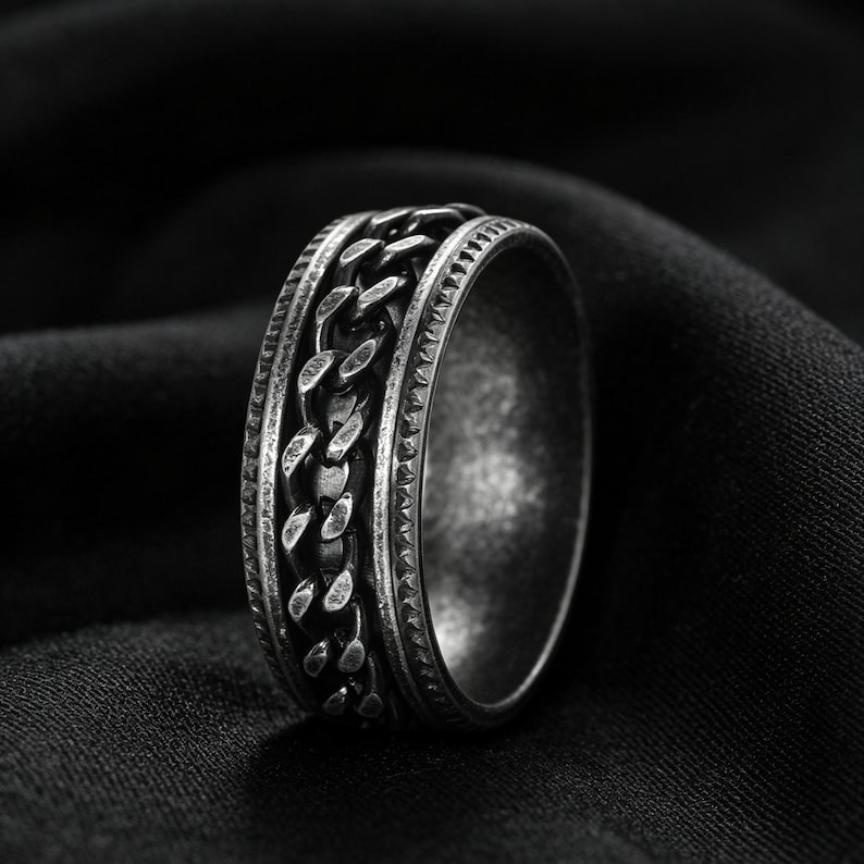 Peut inclure: Une bague en m&eacute;tal argent&eacute; dot&eacute;e d'une bande centrale rotative &agrave; maillons de cha&icirc;ne, encadr&eacute;e par deux bords textur&eacute;s avec une finition sombre et oxyd&eacute;e. L'anneau pr&eacute;sente un profil large et robuste &agrave; l'aspect vieilli et d&eacute;taill&eacute;.