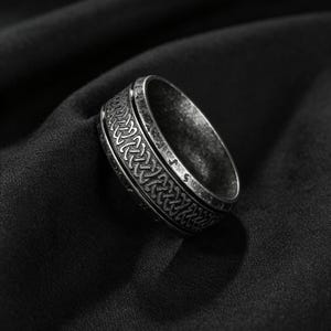 Peut inclure: Une bague en m&eacute;tal argent&eacute; avec une finition sombre et patin&eacute;e, orn&eacute;e d'un motif complexe de n&oelig;uds celtiques r&eacute;p&eacute;t&eacute;s grav&eacute; sur la bande centrale. Les bords de la bague sont lisses et unis, contrastant avec le design textur&eacute; et d&eacute;taill&eacute;.
