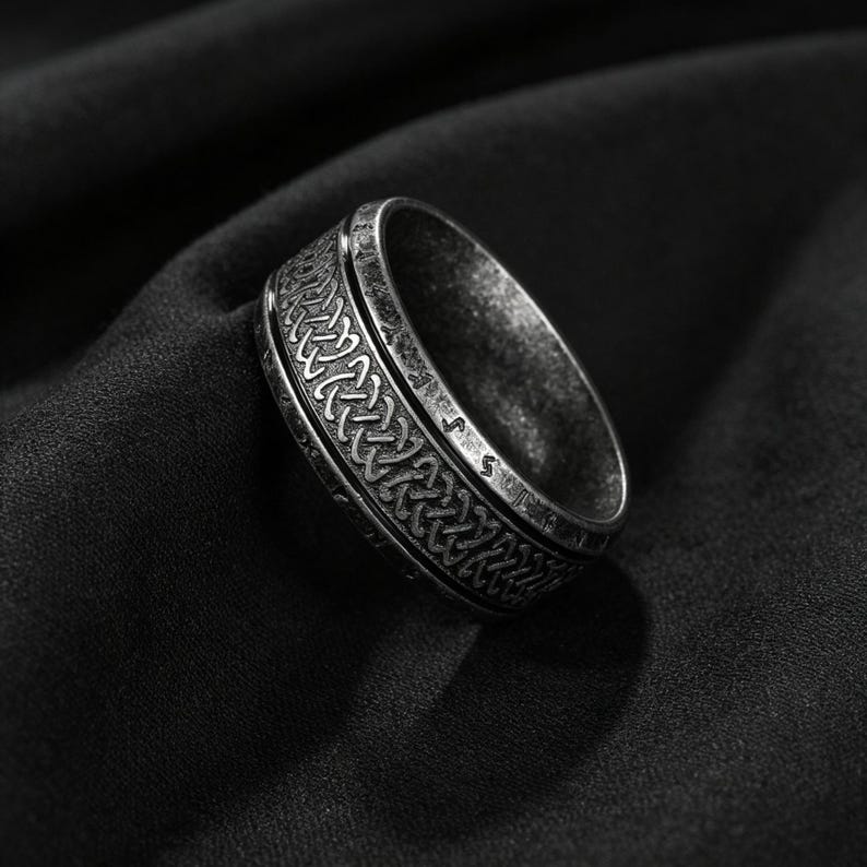 Peut inclure: Une bague en m&eacute;tal argent&eacute; avec une finition sombre et patin&eacute;e, orn&eacute;e d'un motif complexe de n&oelig;uds celtiques r&eacute;p&eacute;t&eacute;s grav&eacute; sur la bande centrale. Les bords de la bague sont lisses et unis, contrastant avec le design textur&eacute; et d&eacute;taill&eacute;.