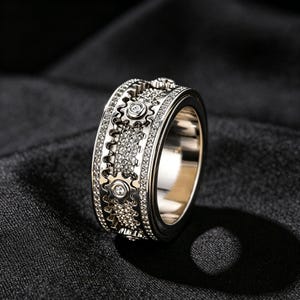 May include: A silver-toned metal ring featuring a wide band with a mechanical gear design. The center of the band is textured with small, sparkling stones and includes several gear-shaped elements, each accented with a single clear stone in the center.