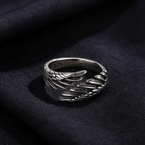 May include: A silver ring with a wing design. The ring has detailed feather patterns and the number "925" is visible. The ring is set against a dark background, highlighting its metallic sheen.
